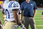 Football coach in navy polo and khaki pants standing on field with players in white and blue uniforms at night.