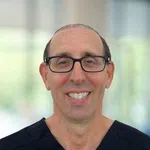 Smiling middle-aged man with glasses wearing black medical scrubs against a blurred background.