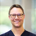Smiling man wearing glasses and dark scrubs with blurred indoor background.