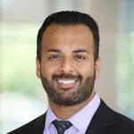 Professional man smiling wearing a black suit, light purple shirt, and striped tie with a blurred background.