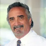 Smiling middle-aged man with gray hair and beard wearing white coat and patterned tie in a bright setting