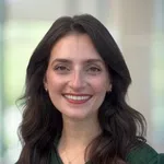 Smiling woman with long dark hair wearing a green top and gold necklace in a bright indoor setting