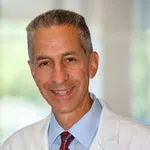 Smiling male doctor wearing white coat, blue shirt, and red paisley tie in bright indoor setting