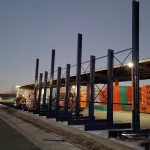 Steel framework under construction in a lumber yard during twilight with stacked wood panels in the background