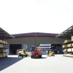 Outdoor lumber yard with stacked wooden planks and forklifts operating in front of a warehouse under clear sky.