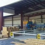 Worker stacking wooden planks inside a spacious industrial warehouse with metal machinery and open roof structure