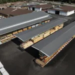 Aerial view of a lumber yard with stacks of wood stored under metal-roofed shelters and adjacent buildings.