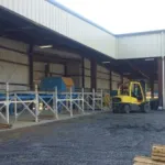 Industrial warehouse exterior with forklift and conveyor system on a gravel surface under metal roofing