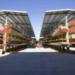 Outdoor lumber yard with organized stacks of wood under metal canopies and a forklift in the center aisle.
