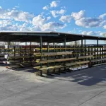 Outdoor lumber storage racks filled with wood planks under a metal roof with a partly cloudy sky.