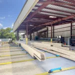 Wood planks stacked under a large metal-roofed warehouse with open sides on a sunny day.