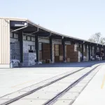 Outdoor warehouse storage with stacks of bundled packages and railway tracks in front under clear sky.