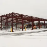 Steel frame structure under construction on a snowy site with construction equipment and cloudy sky