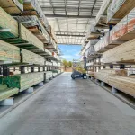Lumber yard aisle with stacked wooden planks on metal racks and a yellow forklift under a metal roof.