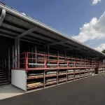 Outdoor warehouse racks with multiple shelves storing wooden planks under a metal roof on a sunny day.