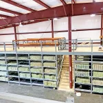Warehouse with stacked wooden planks on metal shelves and a mezzanine level with wooden stairs and railings.
