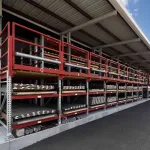 Outdoor industrial storage racks with red shelving holding metal pipes under a roof on a paved area.
