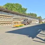 Outdoor lumber yard with stacked wood planks, trucks, forklifts, and clear blue sky on a sunny day.