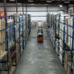 Warehouse aisles with metal racks filled with boxed goods and a forklift in the center aisle under bright lighting.