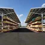 Outdoor lumber storage racks with various wood planks under metal roofs on a clear sunny day with blue sky.