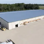 Large industrial warehouse with metal roof, forklift, and stacked wooden pallets in a spacious outdoor yard.