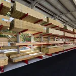 Shelves stocked with stacks of cut lumber under a metal roof in an outdoor storage area on a clear day
