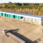 Large industrial warehouse with green doors and a white semi-truck parked in a dirt lot on a sunny day