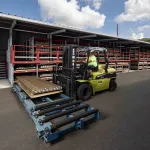 Forklift operator moving metal sheets on roller conveyor outside large warehouse with metal storage racks.