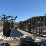 Construction site with steel frame structures and concrete foundation under clear blue sky, surrounded by trees and snow patches.
