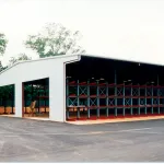 Large open warehouse with metal shelving units and paved driveway under cloudy sky near trees