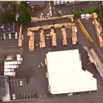 Aerial view of a warehouse with stacked wooden crates, surrounding buildings, parked cars, and nearby train tracks.