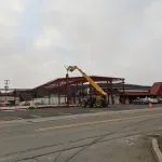 Construction site with steel frame structure and yellow forklift lifting materials on a cloudy day beside a restaurant.