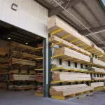 Stacks of lumber neatly organized on yellow racks inside and outside a covered warehouse under a clear sky