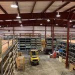 Wide view of a warehouse interior with metal shelving, stocked goods, a yellow forklift, and a pickup truck.