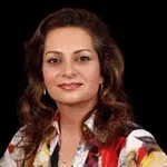 Smiling woman with wavy hair wearing a colorful patterned blouse against a black background