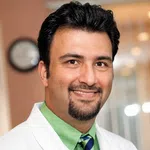 Smiling male doctor in white coat, green shirt, and striped tie in a medical office with a clock in the background.