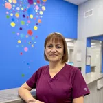 Smiling woman in purple scrubs standing at a counter with colorful circular wall art in the background.