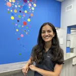 Smiling woman in gray scrubs leaning on counter with colorful dots on blue wall behind her.