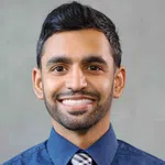 Smiling young man with dark hair and beard wearing a blue shirt and checkered tie against a gray background