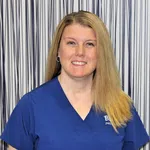Smiling woman with long blonde hair wearing blue medical scrubs standing in front of a vertical striped background.
