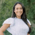 Smiling woman with long dark hair wearing a white dress standing outdoors with greenery background.