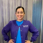 Smiling woman in purple Burleson pediatric dentistry jacket standing with hands on hips in front of striped blue background