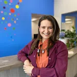 Smiling female dentist wearing maroon scrubs and dental loupes, standing by clinic counter with colorful wall art.