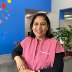 Smiling female dentist in pink scrubs standing by reception desk with colorful blue wall background.