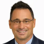 Smiling man with glasses wearing a navy suit and light blue shirt on white background