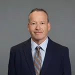Professional middle-aged man in navy suit and striped tie smiling against a plain gray background
