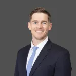 Portrait of a smiling man in a dark suit with a light blue tie against a gray background