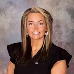 Smiling woman with blonde and brown hair wearing black top with ruffled sleeves and earrings against mottled gray background