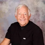 Smiling elderly man with white hair wearing black medical scrubs sitting against a neutral textured background