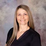 Smiling woman with long straight hair wearing black scrub top against a neutral studio background